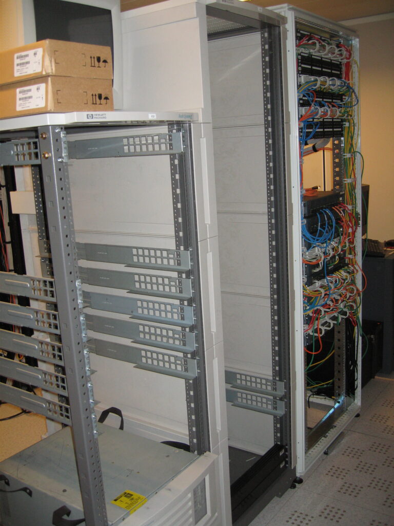 Open equipment racks in a server room, with an empty rack in the center and densely cabled equipment on the right.